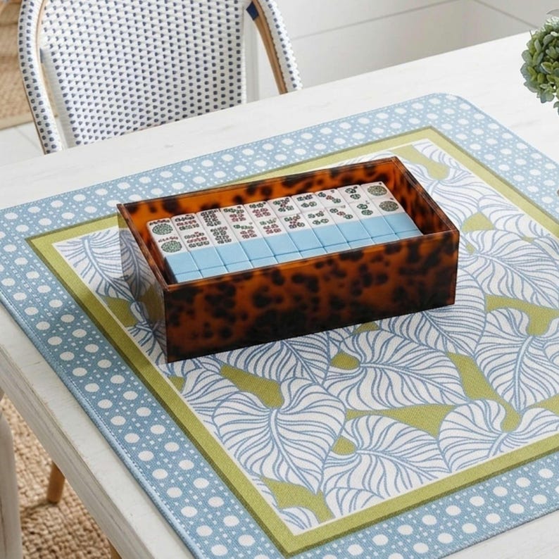 May include: A rectangular box with a brown and black tortoiseshell pattern containing a set of mahjong tiles. The tiles are arranged in rows, featuring white faces with green and red symbols and light blue backs, neatly organized within the container.
