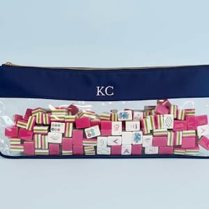 May include: A navy blue zipped pouch with a clear front, filled with pink and white Mahjong tiles. The pouch has gold hardware and the initials "KC" in white. The tiles have green and white stripes and various symbols.