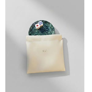 May include: A cream-colored leather pouch with the initials "KC" embossed on the front. A small, round, green and black marble tray with two dice is partially visible. The dice are white with red and blue dots. The pouch is open, revealing the tray.