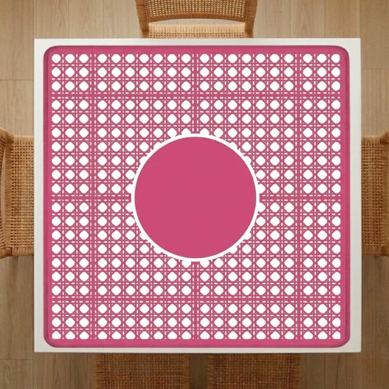 Pu&ograve; includere: Vista dall'alto di un tavolo quadrato con cornice bianca e motivo intrecciato rosa. Il centro presenta un cerchio rosa pieno. Sedie in vimini parzialmente visibili, che suggeriscono un ambiente da pranzo o all'aperto.