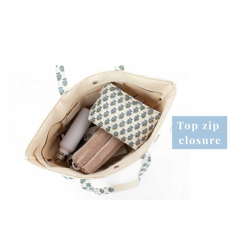 May include: Cream tote bag with blue floral pattern on handles and trim. The bag is open, showing a water bottle, patterned pouch, and a soft, light brown case. Text reads "Top zip closure".