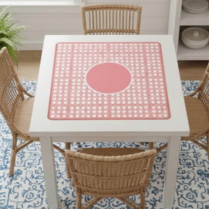 Pu&ograve; includere: Un tavolo quadrato bianco con un sottopiatto rosa e bianco a fantasia. Il sottopiatto ha un cerchio centrale rosa e un bordo bianco smerlato. Quattro sedie in vimini circondano il tavolo e sotto c'&egrave; un tappeto a fantasia blu e bianco.