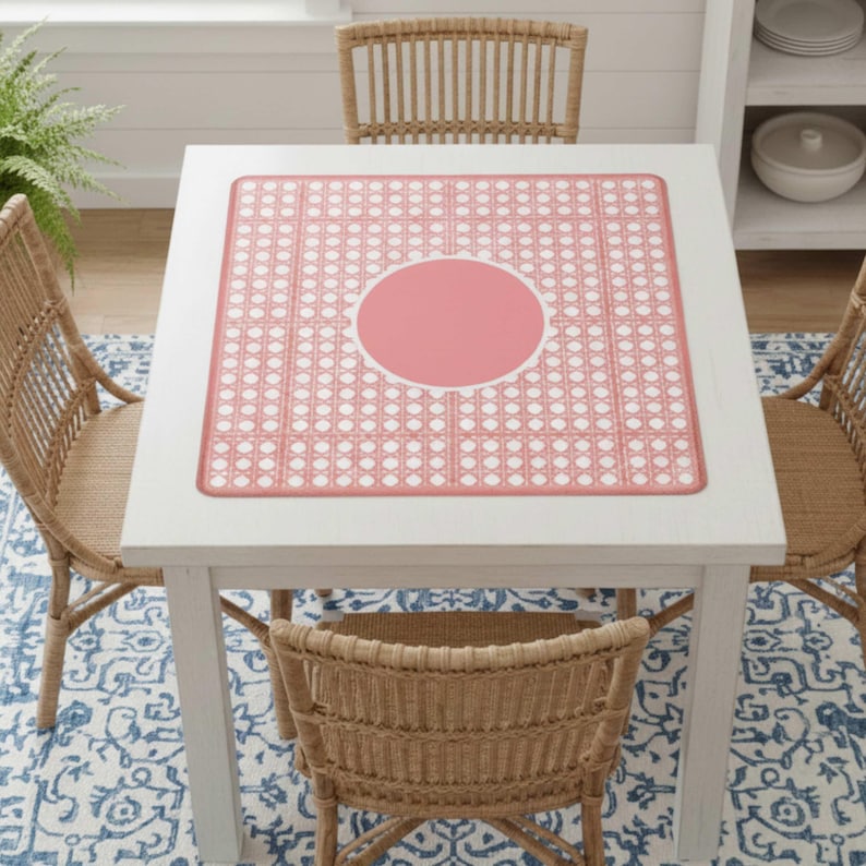 Pu&ograve; includere: Un tavolo quadrato bianco con un sottopiatto rosa e bianco a fantasia. Il sottopiatto ha un cerchio centrale rosa e un bordo bianco smerlato. Quattro sedie in vimini circondano il tavolo e sotto c'&egrave; un tappeto a fantasia blu e bianco.
