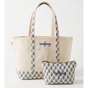 May include: A cream-coloured tote bag and matching pouch set. The tote features a navy blue "mahjong" script, with floral patterned accents on the handles and base. The pouch has a similar floral pattern and "mahjong" script.