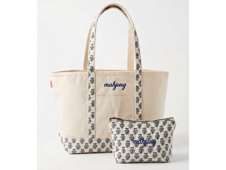 May include: A cream-coloured tote bag and matching pouch set. The tote features a navy blue "mahjong" script, with floral patterned accents on the handles and base. The pouch has a similar floral pattern and "mahjong" script.