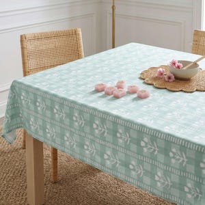 May include: A light green and white floral tablecloth covers a wooden table. The tablecloth has a checkered pattern with floral accents. Pink candles and a bowl with flowers sit on the table. Wicker chairs are in the background.