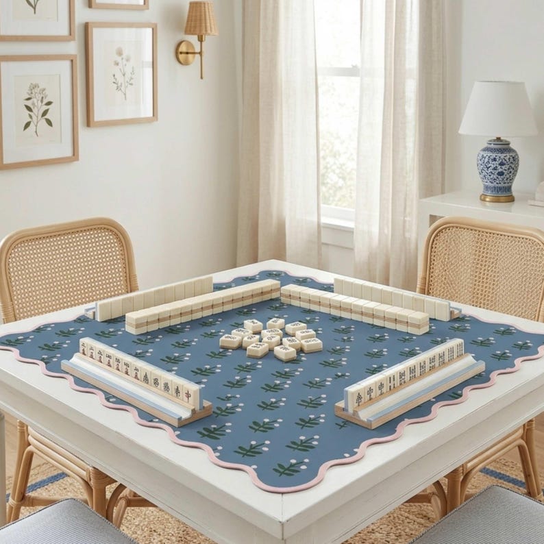 May include: A white square table with a blue floral patterned mat, set for a game of Mahjong. The table has a scalloped edge with pink trim. Rattan chairs surround the table, and framed botanical prints hang on the wall.