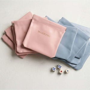 May include: A collection of pink and blue pouches with the word "MAHJONG" printed in gold. The pouches are arranged with a few dice on a white surface. The pouches are made of a soft material.