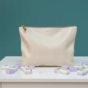 Puede incluir: Una bolsa con cremallera de color crema con un tirador de cremallera estilo bambú. La bolsa está sobre una superficie blanca con varias fichas de Mahjong en tonos morados, verdes y blancos esparcidas alrededor. La bolsa tiene las iniciales "abc".