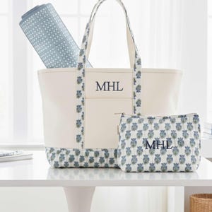 May include: Cream-colored tote bag and matching cosmetic bag with blue floral pattern accents. The tote has a pocket and the initials "MHL". A rolled-up blue yoga mat is inside the tote. The set is on a white table.