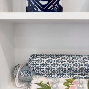 May include: Three cylindrical fabric rolls with white rope drawstrings. The top roll features a blue and white leaf pattern. The middle roll has a light blue floral print. The bottom roll displays a white fabric with green leaves and pink flowers.