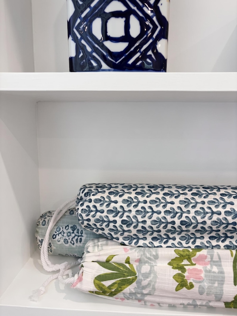 May include: Three cylindrical fabric rolls with white rope drawstrings. The top roll features a blue and white leaf pattern. The middle roll has a light blue floral print. The bottom roll displays a white fabric with green leaves and pink flowers.