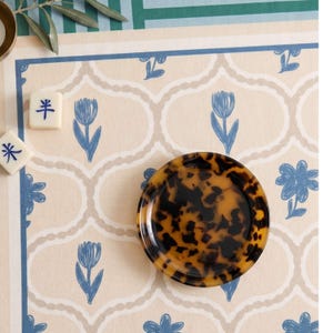 May include: A small, round, tortoiseshell-patterned dish sits on a patterned surface with blue floral and geometric designs. Two Mahjong tiles are visible. The background features a beige and blue color scheme.