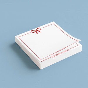 以下が含まれることがあります： 赤いリボンのグラフィックと赤いボーダーが付いた白いメモ帳の束。下部には「KATHERINE SMITH」の文字が赤色で印刷されています。背景は無地の水色です。一番上のシートが少し持ち上がっています。