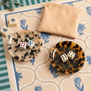 May include: A collection of game pieces including dice, small decorative trays, and a leather pouch. The trays have a tortoiseshell pattern and hold dice with red and blue dots. The pouch is a light tan color and has the initials "CK" embossed on it.