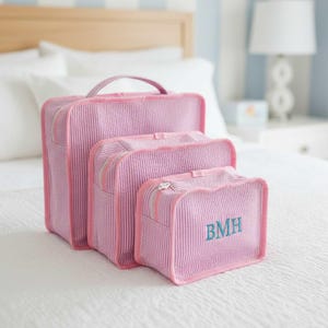 May include: Three pink, vertically striped travel bags of varying sizes are arranged on a white bed. The smallest bag has the initials "BMH" embroidered in teal. Each bag has a zipper closure and a handle.