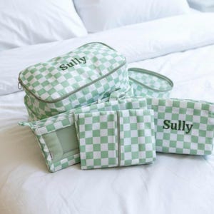 May include: A collection of matching travel bags and pouches in a light green and white checkered pattern. The bags are various sizes and shapes, with the name "Sully" printed on some. The set is arranged on a white bedspread.