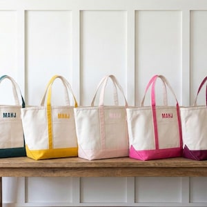 May include: Five canvas tote bags with different colored accents: teal, yellow, pink, magenta, and burgundy. Each bag has a matching base and handles, with the initials "MAHJ" printed on the front. The bags are displayed on a wooden table.