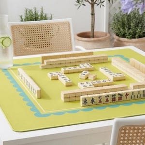 K&ouml;nnte beinhalten: Ein Mahjong-Spiel auf einer gr&uuml;nen Matte mit blauem Wellenrand. Die Spielsteine sind beige mit schwarzen Zeichen. Ein Lautsprecher im Rattan-Stil und ein Glas Wasser mit Gurkenscheiben befinden sich im Hintergrund. Das Spiel steht auf einem wei&szlig;en Tisch.