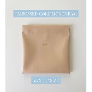 May include: A beige square pouch with the embossed gold monogram "CK". The text "EMBOSSED GOLD MONOGRAM" and "3.5 X 3.5" SIZE" are in light blue rectangles above and below the pouch. The pouch is 3.5 inches by 3.5 inches.