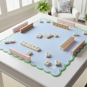 Könnte beinhalten: Ein hellblaues Mahjong-Spielset auf einem weißen Tisch. Das Set enthält Spielsteine, rosa- und beigefarbene Spielsteinhalter und eine blaue Spielfläche mit einem grünen, wellenförmigen Rand. Die Spielsteine sind beige mit schwarzen und roten Symbolen.