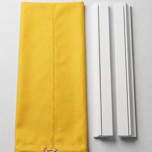 May include: A rectangular mustard yellow fabric panel with a vertical seam and a bottom hem featuring a metal grommet and a white drawstring cord, positioned next to two separate white wooden mounting rails with vertical grooves.