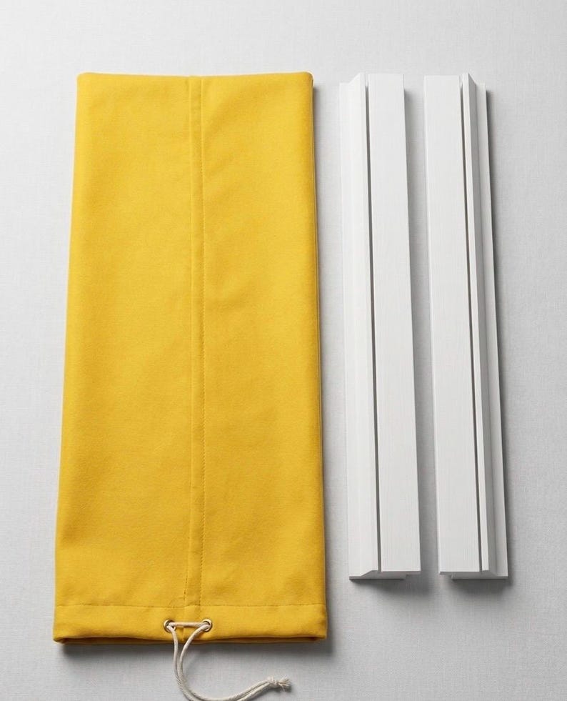 May include: A rectangular mustard yellow fabric panel with a vertical seam and a bottom hem featuring a metal grommet and a white drawstring cord, positioned next to two separate white wooden mounting rails with vertical grooves.