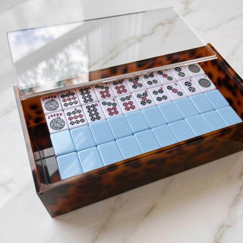 May include: A rectangular box with a brown and black tortoiseshell pattern and a clear acrylic lid. Inside, rows of Mahjong tiles are arranged, featuring white tiles with red and green circular symbols and text reading JOKER, alongside solid light blue tiles.