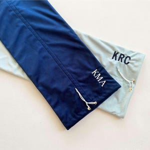 May include: Two rectangular fabric drawstring bags, one in dark navy blue with white KMA lettering and one in light grey with dark navy blue KRC lettering. Each bag features a white drawstring cord with gold-toned metal end caps for closure.