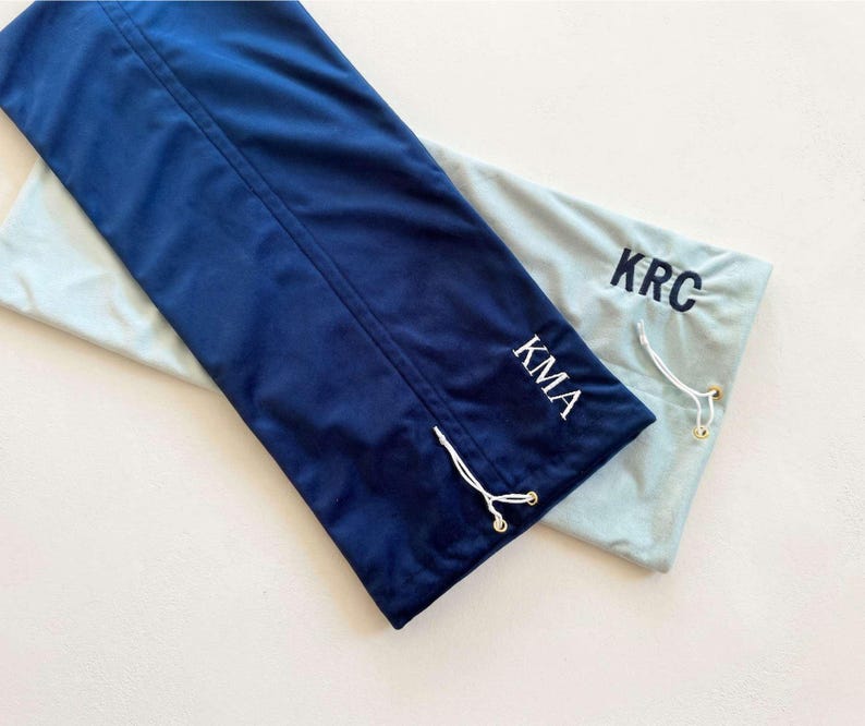 May include: Two rectangular fabric drawstring bags, one in dark navy blue with white KMA lettering and one in light grey with dark navy blue KRC lettering. Each bag features a white drawstring cord with gold-toned metal end caps for closure.