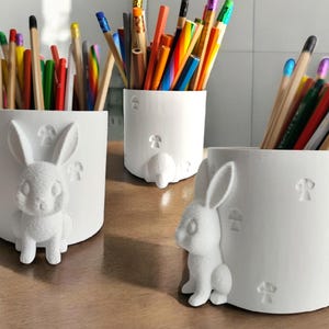 Könnte beinhalten: Drei weiße Stifthalter mit Hasen- und Pilzdesign. Jeder zylindrische Halter ist mit farbigen Bleistiften gefüllt. Der Hase ist 3D und an der Seite befestigt. Die Halter stehen auf einer Holzoberfläche.