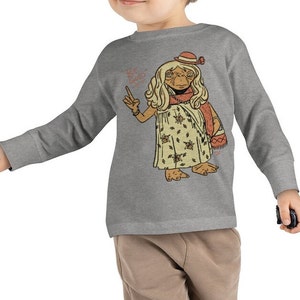 May include: A heather grey long-sleeved t-shirt for children featuring a cartoon illustration of a character from the film E.T. The Extra-Terrestrial. The character is wearing a flower-patterned dress, a hat, and a scarf. The text "Be Kind" is written above the character.