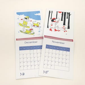 May include: Two spiral-bound wall calendars showing the months of November and December. Each page features a colourful illustration of a person and a dog in seasonal settings, with a grid calendar below the artwork and a small dog icon at the bottom.