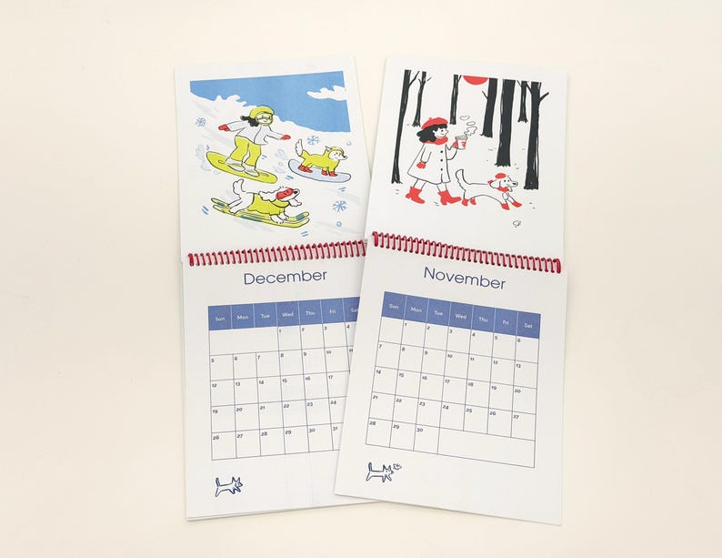 May include: Two spiral-bound wall calendars showing the months of November and December. Each page features a colourful illustration of a person and a dog in seasonal settings, with a grid calendar below the artwork and a small dog icon at the bottom.