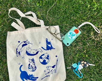 May include: A beige canvas tote bag featuring a blue graphic print of various cats, stars, and the text @vooArt. Resting on the bag are a pair of wire-rimmed glasses, a light green rectangular device with a wrist strap, and a set of metal keys.