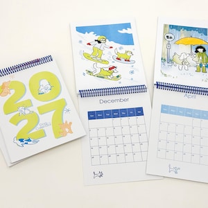 May include: A 2027 spiral-bound dog calendar featuring large lime green numbers on the cover. Inside pages display monthly grids for December and April, accompanied by colourful illustrations of people and dogs in various seasonal settings.
