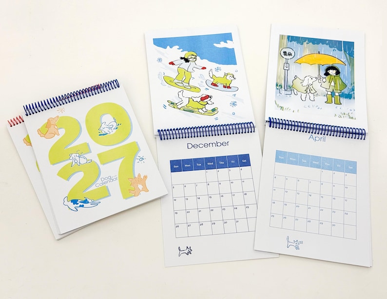 May include: A 2027 spiral-bound dog calendar featuring large lime green numbers on the cover. Inside pages display monthly grids for December and April, accompanied by colourful illustrations of people and dogs in various seasonal settings.