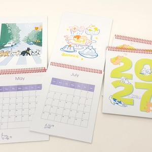 May include: A 2027 dog calendar with spiral binding, featuring monthly grid pages for May and July. The pages display colourful illustrations of dogs in various settings, including a street crossing and summer treats, with the text 2027 Dog Calendar.