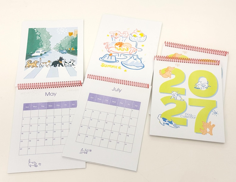 May include: A 2027 dog calendar with spiral binding, featuring monthly grid pages for May and July. The pages display colourful illustrations of dogs in various settings, including a street crossing and summer treats, with the text 2027 Dog Calendar.