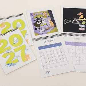 May include: Three 2027 dog calendars with spiral bindings. The covers feature large yellow numbers and small dog illustrations. Open pages show a calendar grid for October and June, with colourful cartoon illustrations of dogs in various settings.