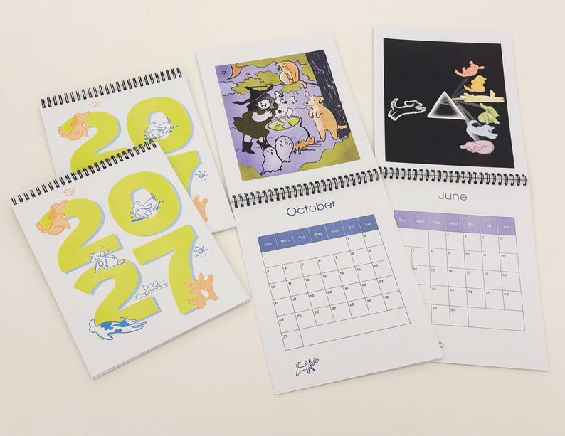 May include: Three 2027 dog calendars with spiral bindings. The covers feature large yellow numbers and small dog illustrations. Open pages show a calendar grid for October and June, with colourful cartoon illustrations of dogs in various settings.