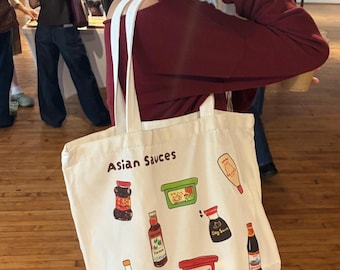 May include: A white canvas tote bag featuring a colourful graphic print of various Asian sauce bottles and containers. The text "Asian Sauces" is printed at the top, and "@UooArt" is printed at the bottom. The bag has two white fabric handles.