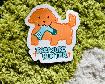 May include: A sticker featuring a cartoon illustration of a light orange dog with a blue sock in its mouth. The text "TREASURE HUNTER" is written in blue and white below the dog.