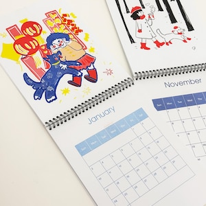 May include: A spiral-bound wall calendar featuring two open pages. The left page shows a January calendar with a colourful illustration of a person and a blue dog near red lanterns. The right page shows a November calendar with a black and white illustration of a person and a dog in a forest.