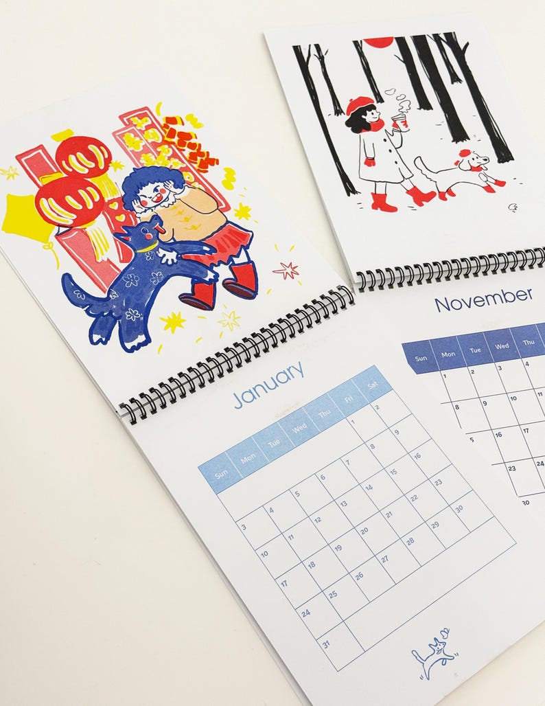 May include: A spiral-bound wall calendar featuring two open pages. The left page shows a January calendar with a colourful illustration of a person and a blue dog near red lanterns. The right page shows a November calendar with a black and white illustration of a person and a dog in a forest.