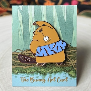 May include: An enamel pin featuring a brown and orange beaver wearing a blue and white striped life preserver. The beaver is smiling and has a small tail. The pin is set against a green and brown background with the text "The Bunny Art Cart".