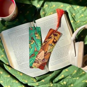 May include: Two illustrated bookmarks with tassels lie on an open book. One bookmark is green with a cartoon turtle wearing a witch hat. The other is orange with a cartoon scene of a ghost and pumpkins. A cup and headphones are also visible.