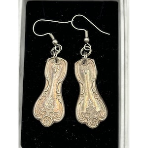 May include: Silver-toned earrings crafted from repurposed silverware. Each earring features a floral design at the base and along the edges, with a decorative pattern. The earrings hang from silver-colored ear wires, presented against a black background.