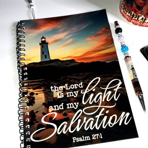 以下が含まれることがあります： 灯台のシーンと「the Lord is my light and my Salvation」というテキストが書かれたスパイラル綴じノート。装飾的なペン、付箋、マグカップも表示されています。