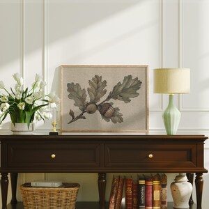 May include: A framed botanical print featuring an oak branch with leaves and acorns. The artwork is displayed on a dark brown console table, complemented by a green lamp, white tulips, and a decorative basket. The print has a neutral color palette.